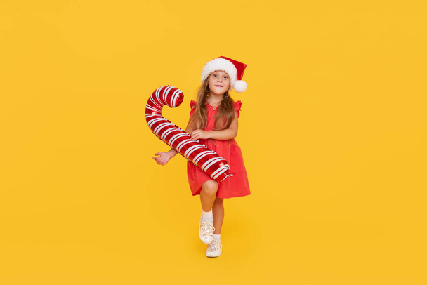 A cute little girl child in a fancy dress and a Santa hat holds an inflatable shape of a candy cane on a yellow background. 2021 new year holidays concept and childrens new year gifts advertising - Foto, imagen