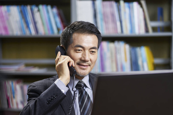 asian corporate executive sitting at desk in office conversing on phone - Photo, Image