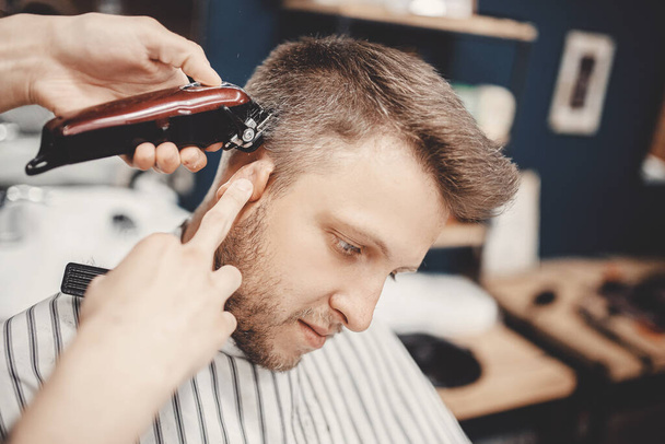 Парикмахер в парикмахерской стрижет волосы электрическим для молодого парня для модной прически - Фото, изображение