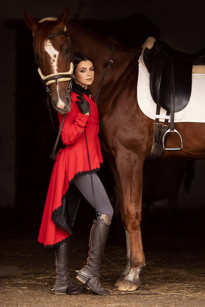 Улыбающаяся жокей женщина стоит с коричневой лошадью в конюшне, черный фон - Фото, изображение