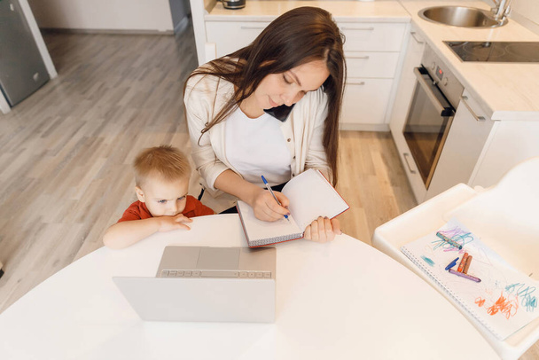 Kobieta na urlopie macierzyńskim pracuje w domu online z laptopem w kuchni z małym dzieckiem. Concept mama praca podczas kwarantanny izolacji podczas Covid-19 - Zdjęcie, obraz