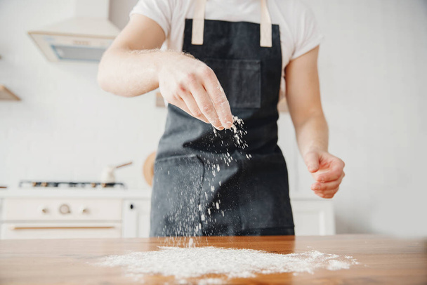 Chef in apron sprinkles flour with hand on dough, wooden table, light background - Photo, Image