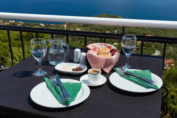 Mesa preparada para dos personas en la terraza del restaurante con vistas a la costa marítima de la isla de Corfú en Grecia. En la mesa café, agua y pasta de aceitunas con pan - Foto, imagen