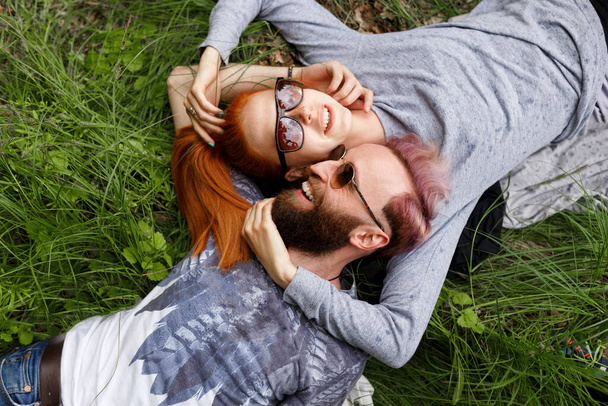 Vista superior de una hermosa pareja, sentada en una hierba verde, sentada con las cabezas giradas usando gafas de sol. - Foto, imagen