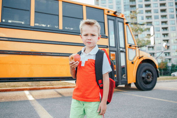 Caucasian boy student with backpack near yellow bus on first September day. Child kid eating apple fruit at school yard outdoor. Education and back to school in Autumn Fall.  - Photo, Image