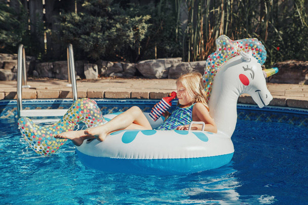 Słodka urocza dziewczyna w okularach przeciwsłonecznych z napojem leżącym na nadmuchiwanym pierścieniu jednorożca. Dziecko bawiące się w basenie. Letnia aktywność wody na świeżym powietrzu dla dzieci.  - Zdjęcie, obraz