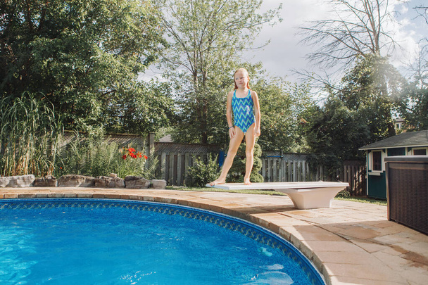 Aranyos vörös hajú lány ugródeszkáról ugrik a medencébe. Gyerek gyerek élvezi az otthoni uszodát. Nyári szabadtéri vízi aktivitás gyerekeknek.  - Fotó, kép