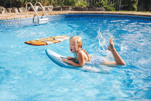 Cute adorable red-haired girl with freckles swimming in pool on swim board. Kid child enjoying having fun in swimming pool. Summer outdoor water activity for kids.  - Photo, Image