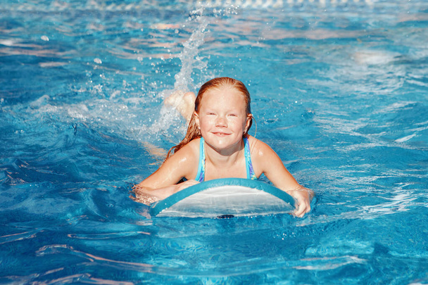 Aranyos, imádnivaló, vörös hajú lány szeplőkkel úszva a medencében az úszódeszkán. Gyerek gyermek élvezi az uszodát. Nyári szabadtéri vízi aktivitás gyerekeknek.  - Fotó, kép