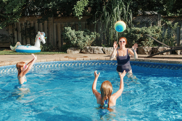 Mother playing ball with daughters children in swimming pool on home backyard. Mom and sisters siblings having fun in swimming pool together. Summer outdoor water activity for family and kids.  - Fotoğraf, Görsel