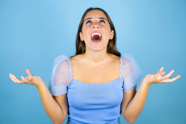 Young beautiful woman over isolated blue background crazy and mad shouting and yelling with aggressive expression and arms raised - Photo, Image
