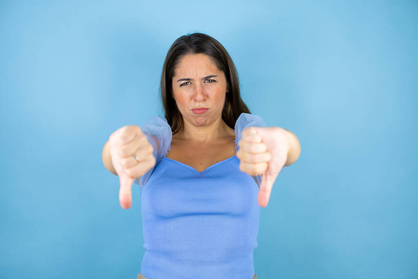Mladá krásná žena nad izolovaným modrým pozadím s rozzlobenou tváří, negativní znamení ukazující odpor s palci dolů - Fotografie, Obrázek