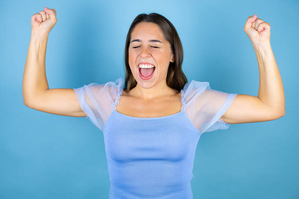 Молодая красивая женщина на изолированном голубом фоне очень счастлива и взволнована делая жест победителя с поднятыми руками, улыбаясь и крича об успехе. - Фото, изображение