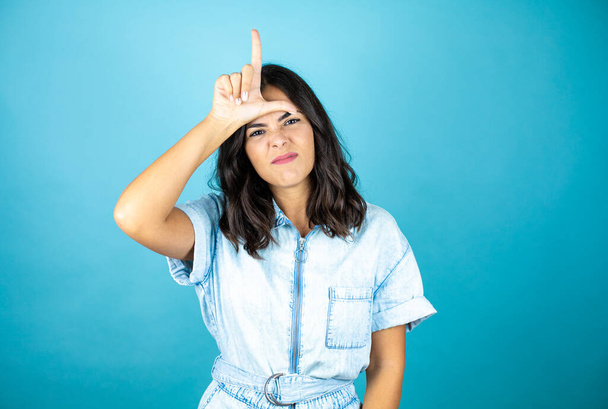 jonge mooie vrouw dragen van een spijkerbroek jumpsuit over geïsoleerde blauwe achtergrond lachen van mensen met vingers op het voorhoofd doen verliezer gebaar spotten en beledigend. - Foto, afbeelding