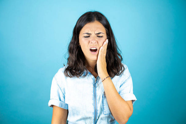 Молода красива жінка в джинсовому комбінезоні над ізольованим синім тлом торкається рота рукою з болючим вираженням через зубний біль або стоматологічні захворювання на зубах
. - Фото, зображення