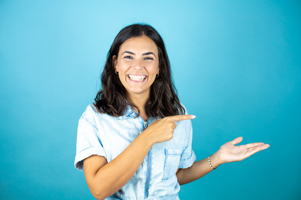 Mladá krásná žena v džínové kombinéze přes izolované modré pozadí s úsměvem, ukazující a ukazující něco, co je na její ruce - Fotografie, Obrázek
