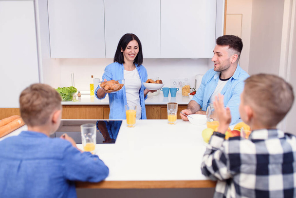 Joyful family of four persons drink orange juice with croissants and tasty cakes in the kitchen. Happy family concept. - Photo, image