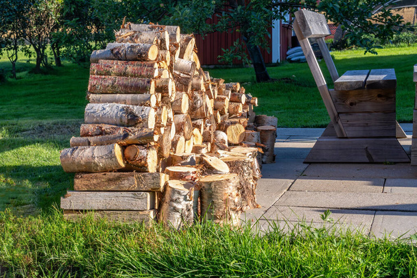 Close up photo of fresh stacked fire wood logs on barbeque spot, garden. A lot of birch logs harvested to be used for grilling. Summer evening, green grass. Blurry apple tree and small wooden cabin - Photo, Image