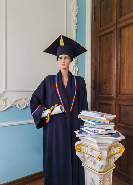 Student mannequin, in een bibliotheek, verkleed als een afgestudeerde met een zwarte academische pet en toga, met een diploma in de hand. Prachtige sierlijke oude zuilen met verschillende boeken. Onderwijsconcept. - Foto, afbeelding