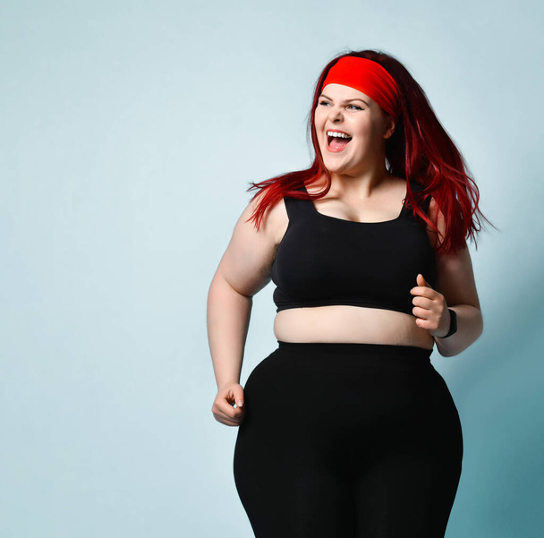 Active young plus-size woman with flowing red hair in black sport top and leggings shouting with laughter and delight on blue background. Copy space - Foto, Imagem