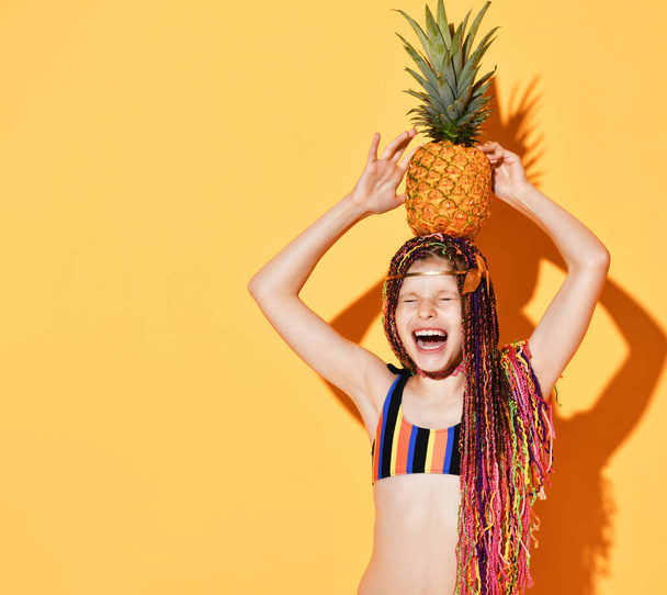 Cheerful girl with colorful pigtails holding a pineapple on her head. - Photo, image