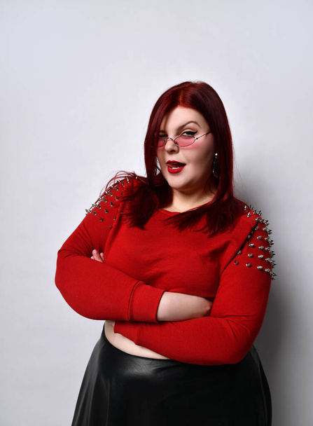 Overweight red-haired woman in a red studded top, black leather skirt and earrings. She stands with her arms folded in a twisted pose - Foto, Bild