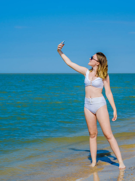 Beautiful young girl on the sea beach near the water posing and taking a selfie on a smartphone. Girl in a white swimsuit and sunglasses. Summer sunny day. Sea vacation concept. Copy space - Φωτογραφία, εικόνα