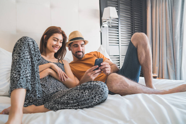Portrait of lovely couple relaxing and using mobile phone while laying on bed at hotel room. Lifestyle and travel concept. - Foto, Bild