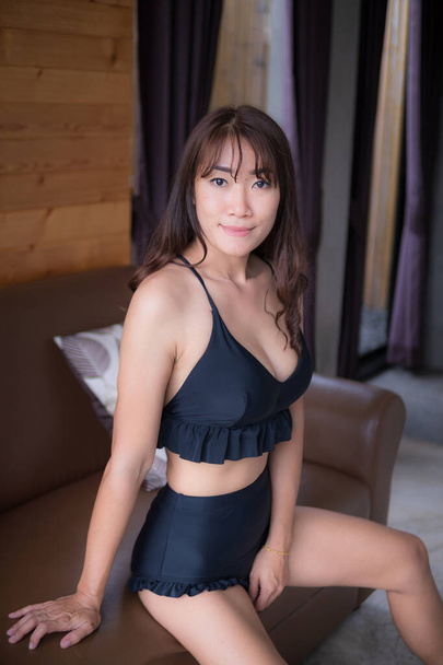 Una donna asiatica in bikini nero sexy si siede sul divano dell'hotel. - Foto, immagini