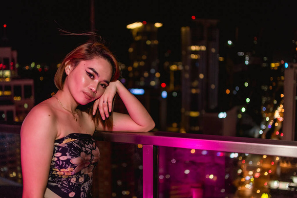 バンダナチューブのトップをした魅力的な若いアジア人女性が街の景色を望む屋上バーでポーズをとっています。マニラ首都圏のマカティで。マニラのナイトライフとデート. - 写真・画像