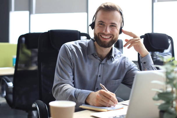 Smiling male call-center operator with headphones sitting at modern office, consulting online information in a laptop, looking up information in a file in order to be of assistance to the client - Photo, Image