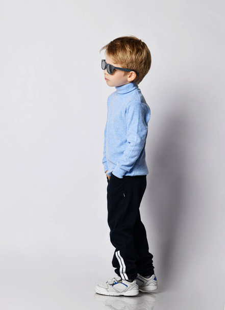 Self-confident blond kid boy in sneakers, black sport pants, blue turtleneck sweater and sunglasses stands side to us - Fotó, kép