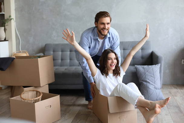 Happy couple is having fun with cardboard boxes in new house at moving day, woman riding sitting in cardboard box while man pushing it. - Photo, Image