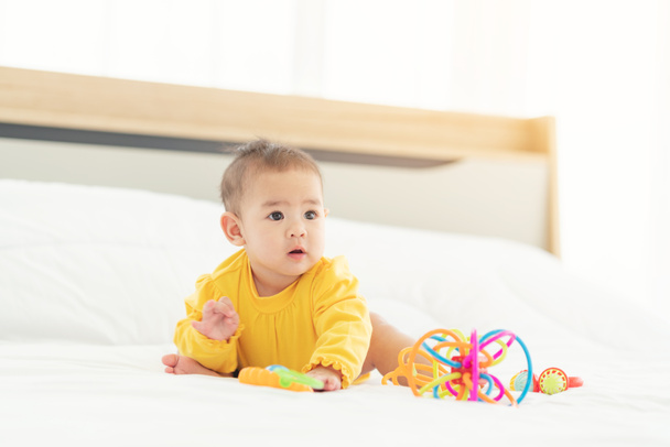 Χαριτωμένο ταϊλανδέζικο μωρό που παίζει περίεργα στο κρεβάτι. Μικρό μωρό με πάνα ξαπλωμένο στην κοιλιά του σε λευκό κρεβάτι με παιχνίδια - Φωτογραφία, εικόνα