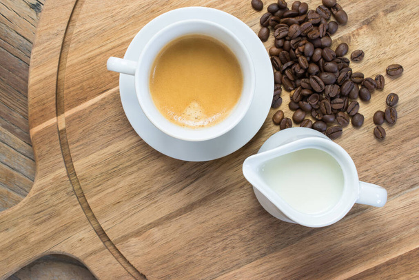 Taza de café cremoso con espuma, jarra de leche y granos de café tostados sobre tabla de madera rústica. Vista superior. - Foto, imagen