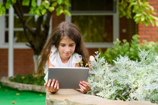 Dziewczyna leży w zielonym ogrodzie i trzyma tabliczkę. Dziewczyna czyta e-booki lub wiadomości w Internecie. - Zdjęcie, obraz