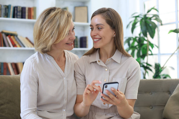 Middle aged mother and adult daughter hugging, using phone together, watching video or photos, sitting on cozy couch at home - Photo, image