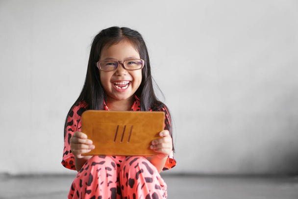 Ασιατικό παιδί φτωχό ή παιδί κορίτσι φορούν μπλε γυαλιά μπλοκ για να μάθουν από το σπίτι ή να μελετήσει σε απευθείας σύνδεση στο tablet του υπολογιστή και την επικοινωνία με χαμόγελο γέλιο ή αστείο παιχνίδι στο σκοτεινό δρόμο και βρώμικο λευκό τοίχο - Φωτογραφία, εικόνα