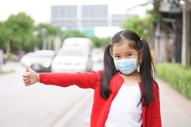 アジアの子供や子供の女の子は、 PM2.5ほこりやコロナウイルスや煙細菌を親指で保護するか、街の通りで道路の車の交通渋滞でヒッチハイクのように病気に近い口の鼻やインフルエンザの顔のマスクを着用 - 写真・画像
