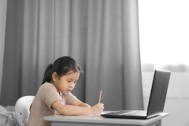 Ασιάτης μαθητής ή παιδί κορίτσι χαμόγελο γράψει ή να σχεδιάσετε και να κάνει την εργασία στο χαρτί με το σημειωματάριο του υπολογιστή για την επικοινωνία και να μελετήσει σε απευθείας σύνδεση ή να μάθουν από το σπίτι για να παίξει φορητό υπολογιστή για πίσω στη σχολική εκπαίδευση - Φωτογραφία, εικόνα