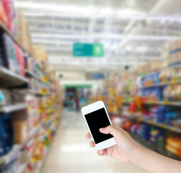hand holding mobile phone with supermarket shelves aisle blurred background - Foto, afbeelding