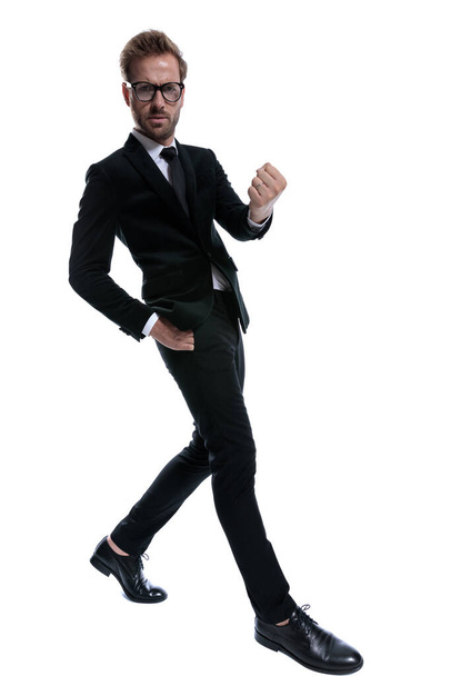 uomo d'affari arrabbiato in abito nero facendo gesto pugno, tenendo la mano in tasca e camminando approssimativamente su sfondo bianco - Foto, immagini