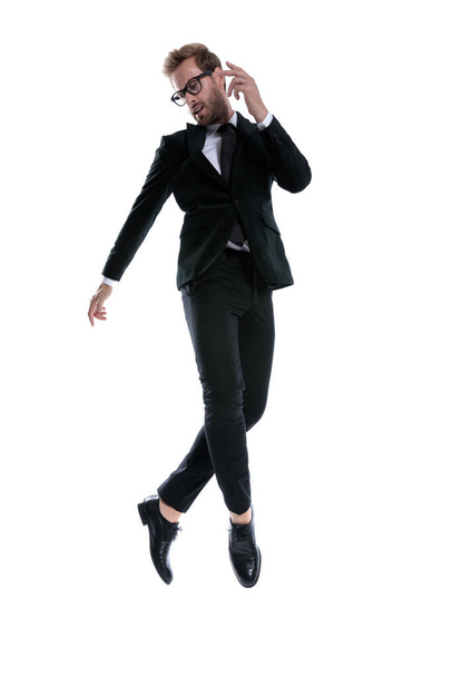 dramático empresario fresco en traje negro mirando hacia abajo lado, saltando en el aire, cruzando las piernas y tomándose de la mano en una pose de moda sobre fondo blanco, cuerpo completo - Foto, Imagen