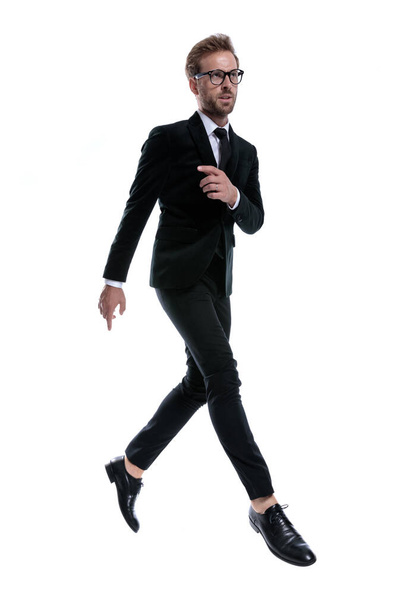 прохладный элегантный мужчина в черном костюме держит руку в позе моды и уверенно прыгает в воздухе и танцует на белом фоне, все тело - Фото, изображение
