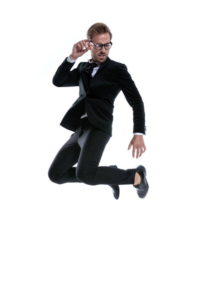 cool homem elegante em terno preto olhando para o lado, fixando óculos e pulando no ar no fundo branco, corpo inteiro - Foto, Imagem