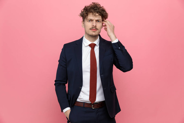 confused young businessman in blue navy suit holding hand to temple and thinking, standing on pink background - Foto, immagini