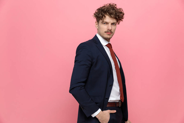 side view of young elegant fashion model in suit holding hands in pockets and walking on pink background - Photo, Image