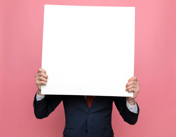 κομψό άνθρωπος της μόδας σε ναυτικό μπλε κοστούμι καλύπτει το πρόσωπο με λευκό άδειο πίνακα και στέκεται σε ροζ φόντο - Φωτογραφία, εικόνα