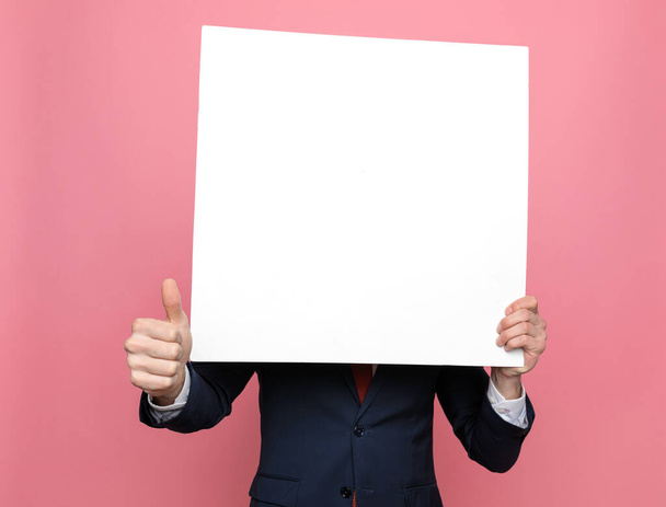 白い空の板の後ろに隠れてピンクの背景に親指を立てるスーツ姿のエレガントなビジネスマン - 写真・画像