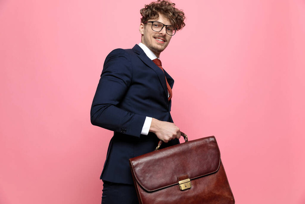 heureux jeune homme d'affaires en costume bleu marine portant des lunettes, tenant une valise, souriant et marchant sur fond rose - Photo, image